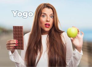 A surprised woman holding a chocolate bar in one hand and a green apple in the other, with the word "Yogo" floating next to her.