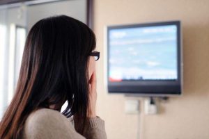 A person with long dark hair and glasses watches a television screen intently, reacting with concern to the negative news being displayed.